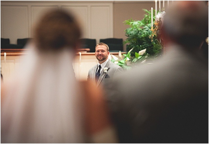 Groom looking at his bride walk down the aisle over the shoulders of the bride and her father during the sparkler exit with a fur wrap during the High Point North Carolina Wedding at First Baptist Church High Point and high point country club at Emerywood