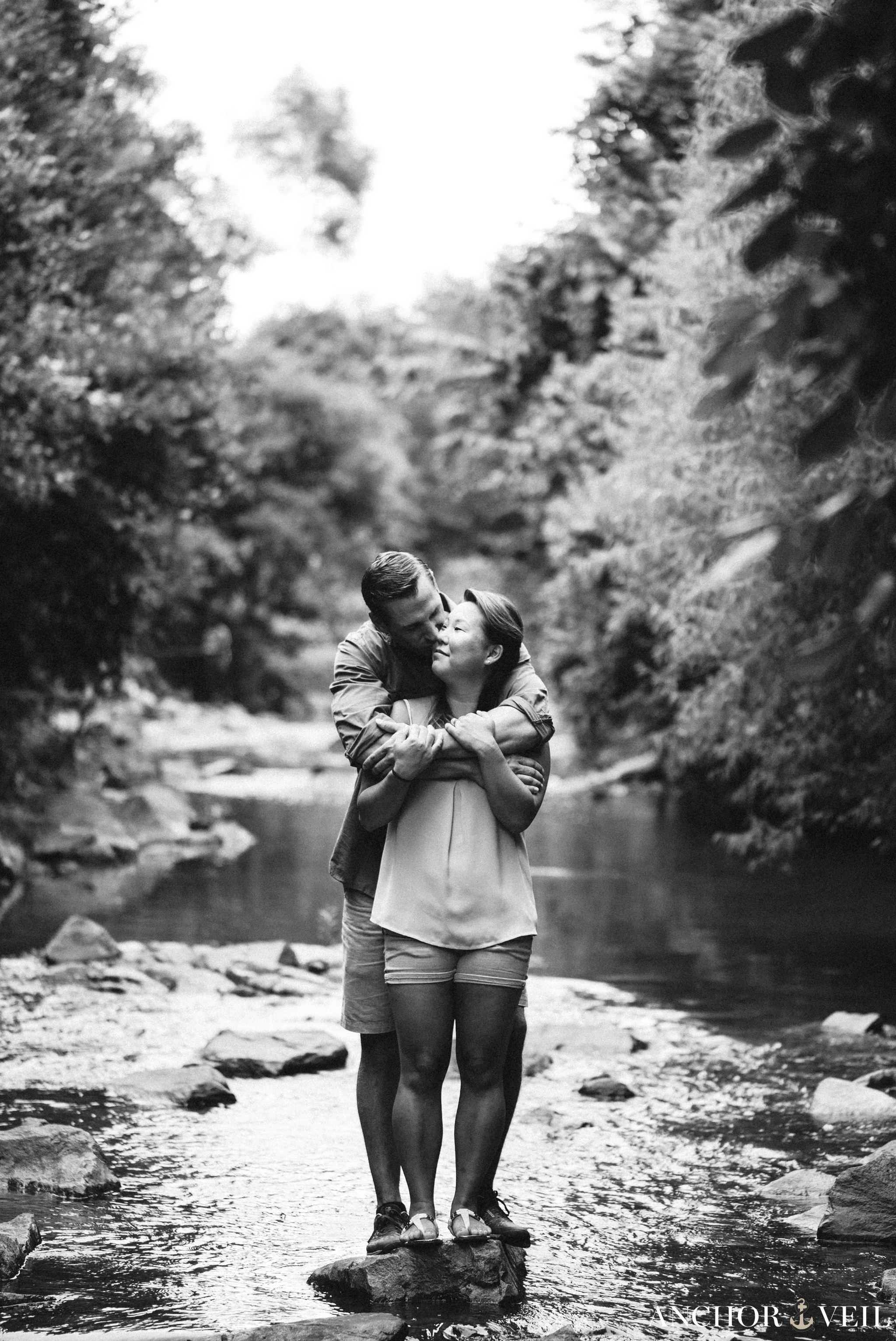 black and white photograph of a lover's tender embrace while standing on the rocks of a creek during their Freedom Park Engagement Session