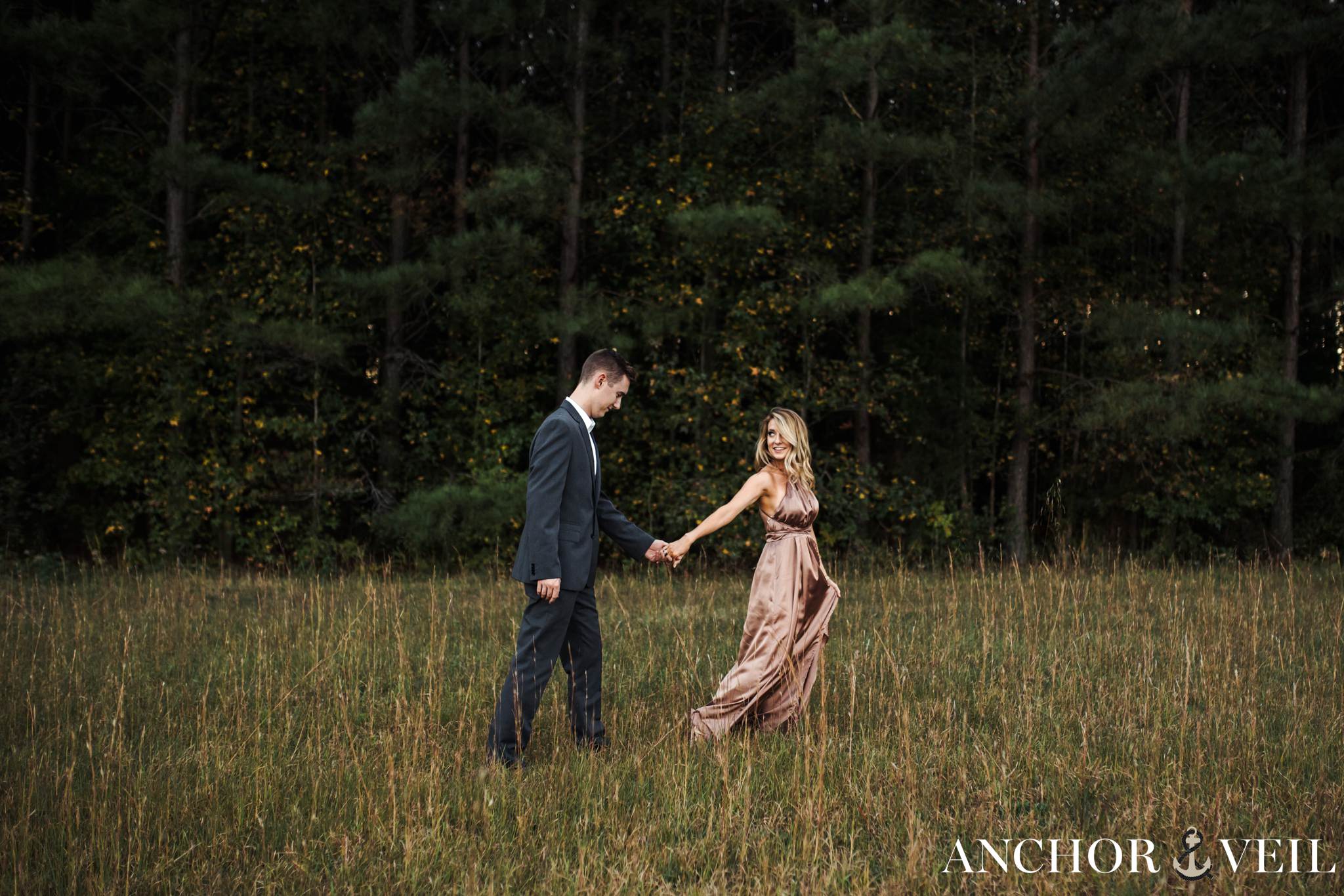 leading him by hand in the field in the tree forest during their charlotte engagement session