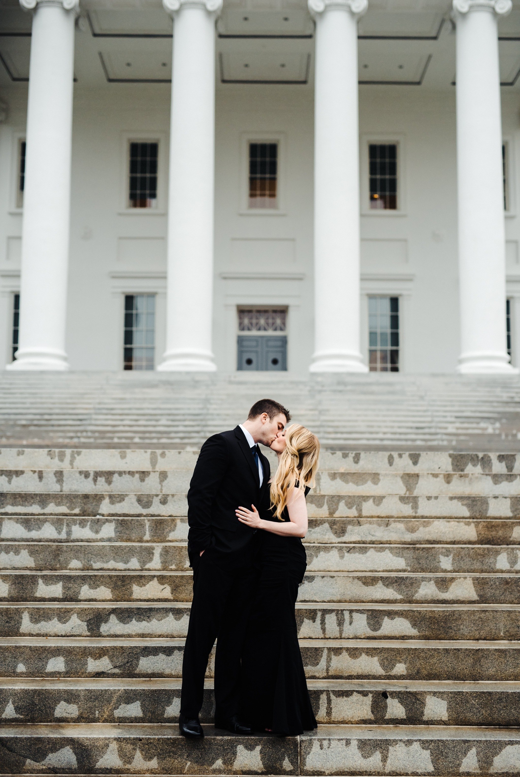 A couple share an intimate kiss with a symmetrical background image of the VA capitol building on their Downtown Richmond Engagement Session