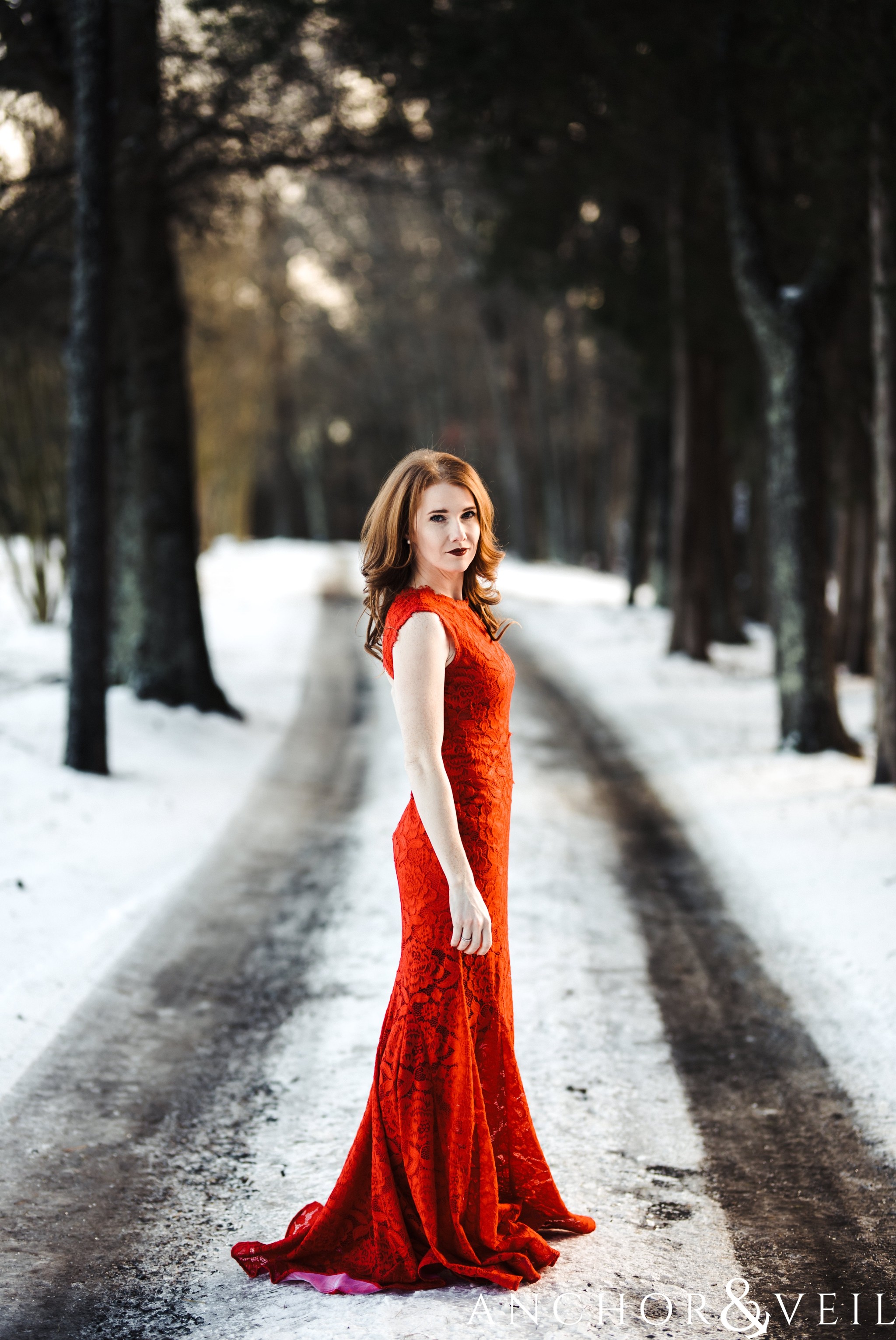 standing in her red dress on the road during their Charlotte Snow engagement session in the woods 