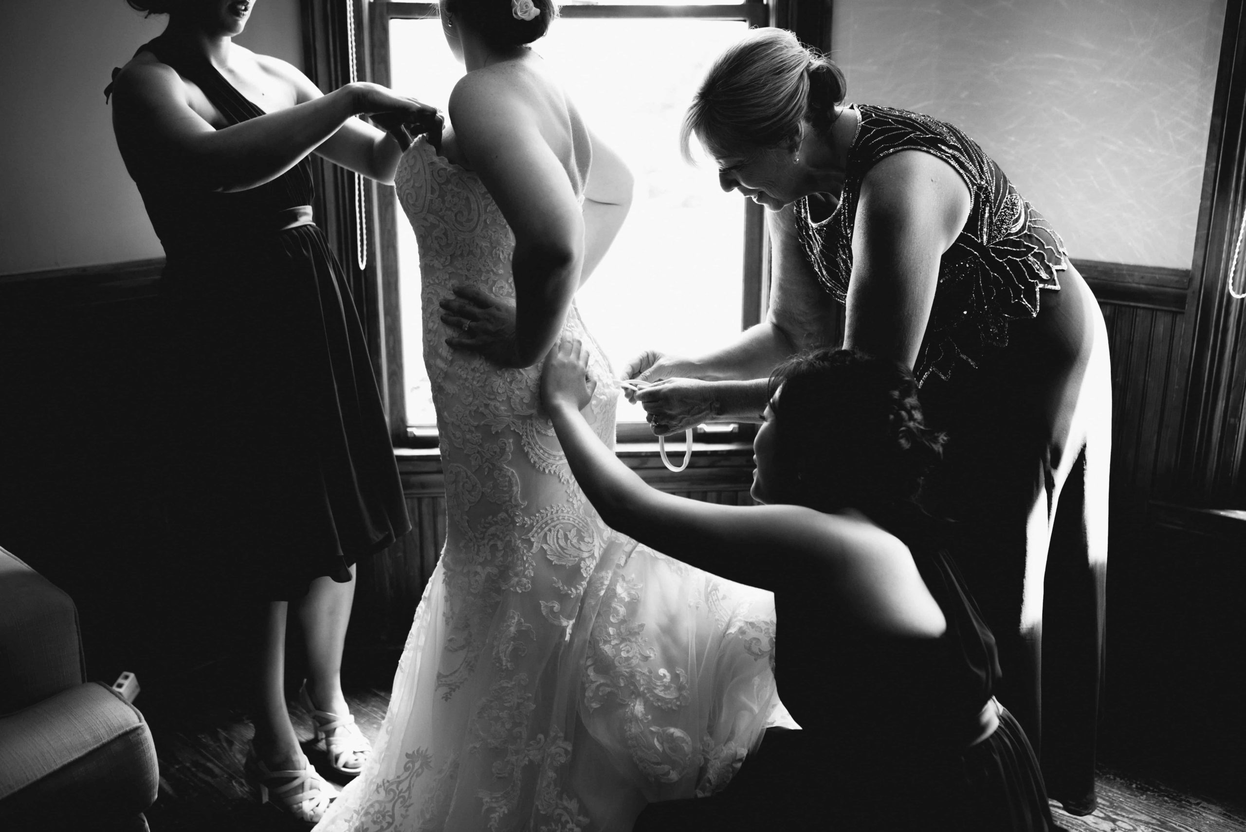 Black and white photography of a bride finished getting her dress on with bridesmaids and mother next to a window at The Whitehead Manor